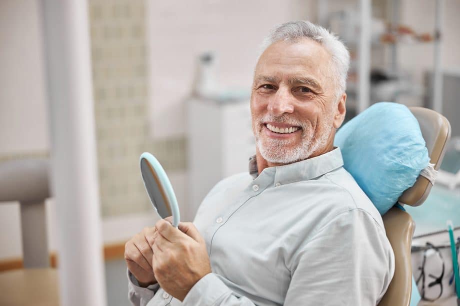 Older man smiling with mirror sitting in dental exam room
